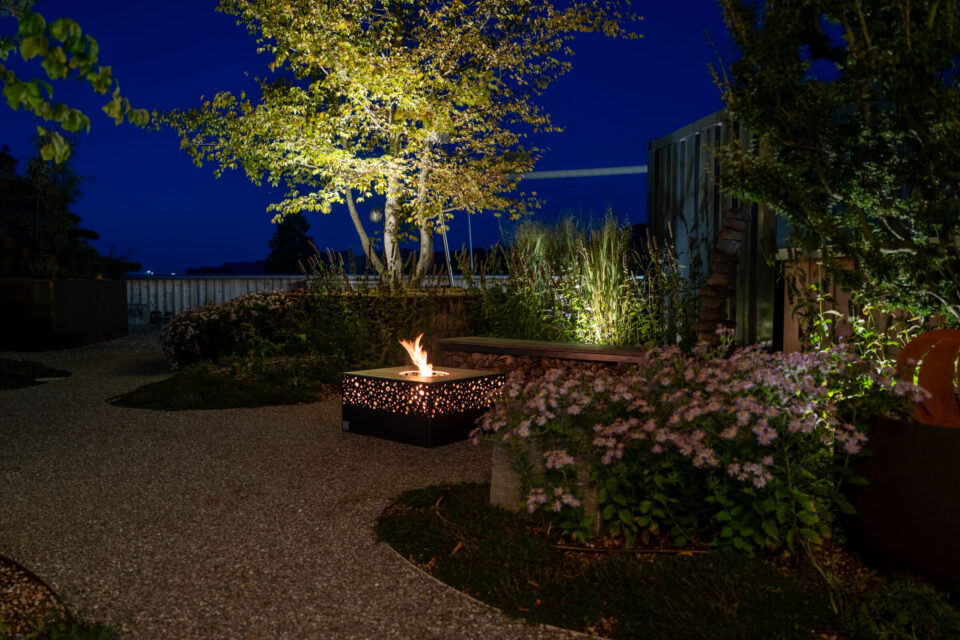 Nächtlicher Garten mit leuchtendem Feuer im Sitzbereich.