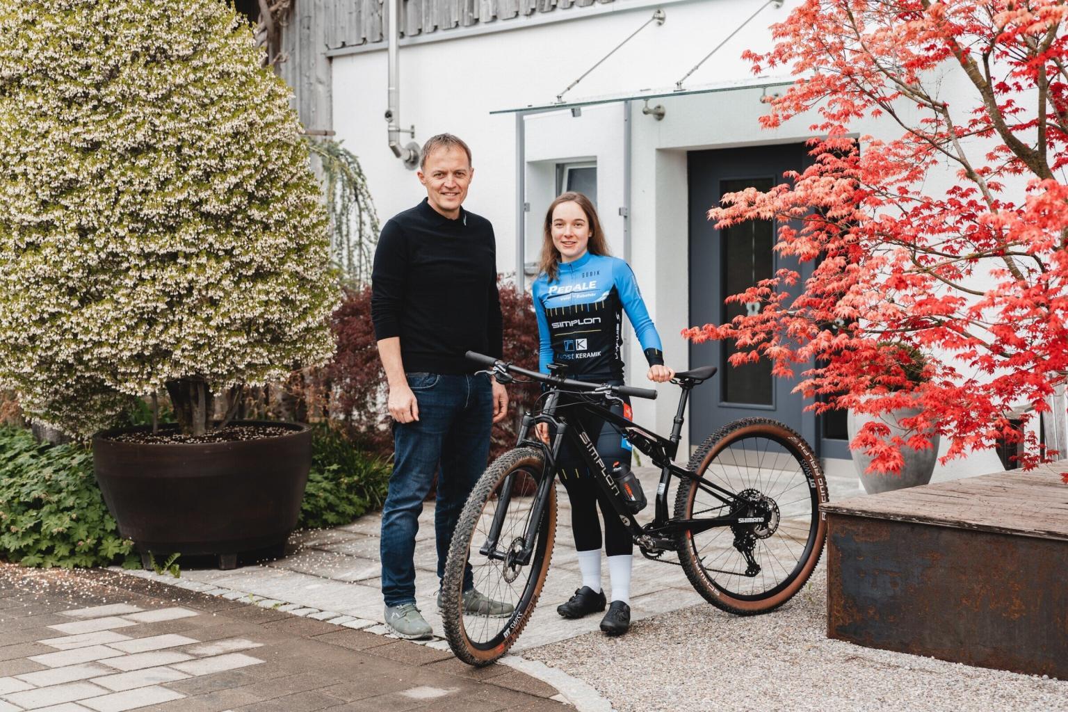 Zwei Personen mit Fahrrad vor Haus.