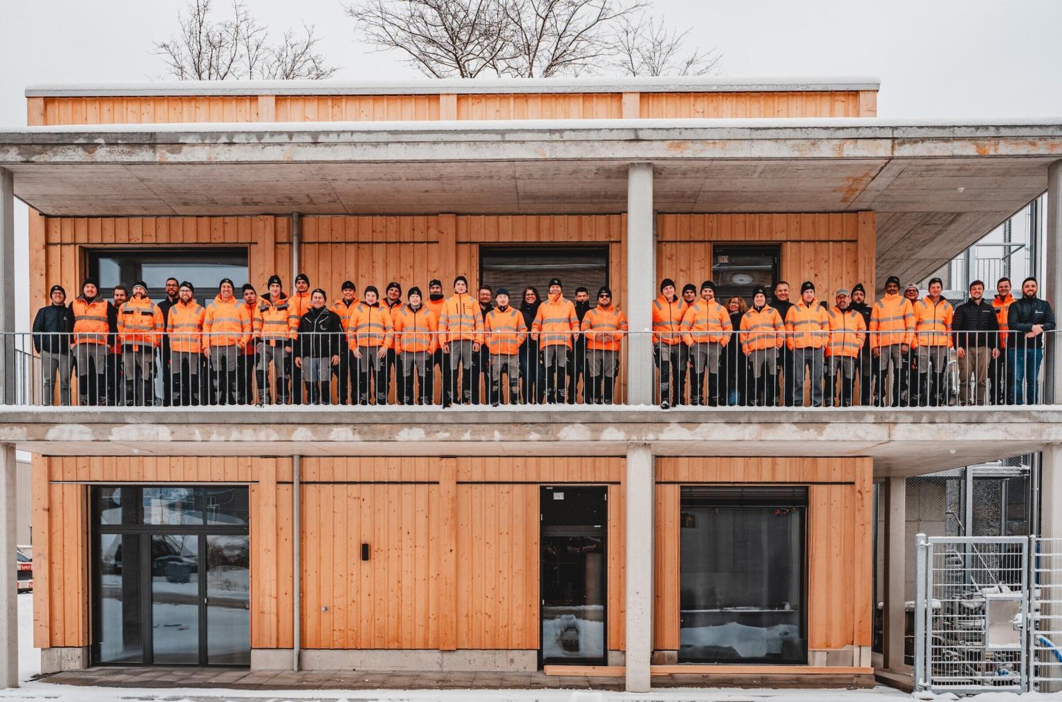 Gruppe in orangen Jacken vor Holzgebäude